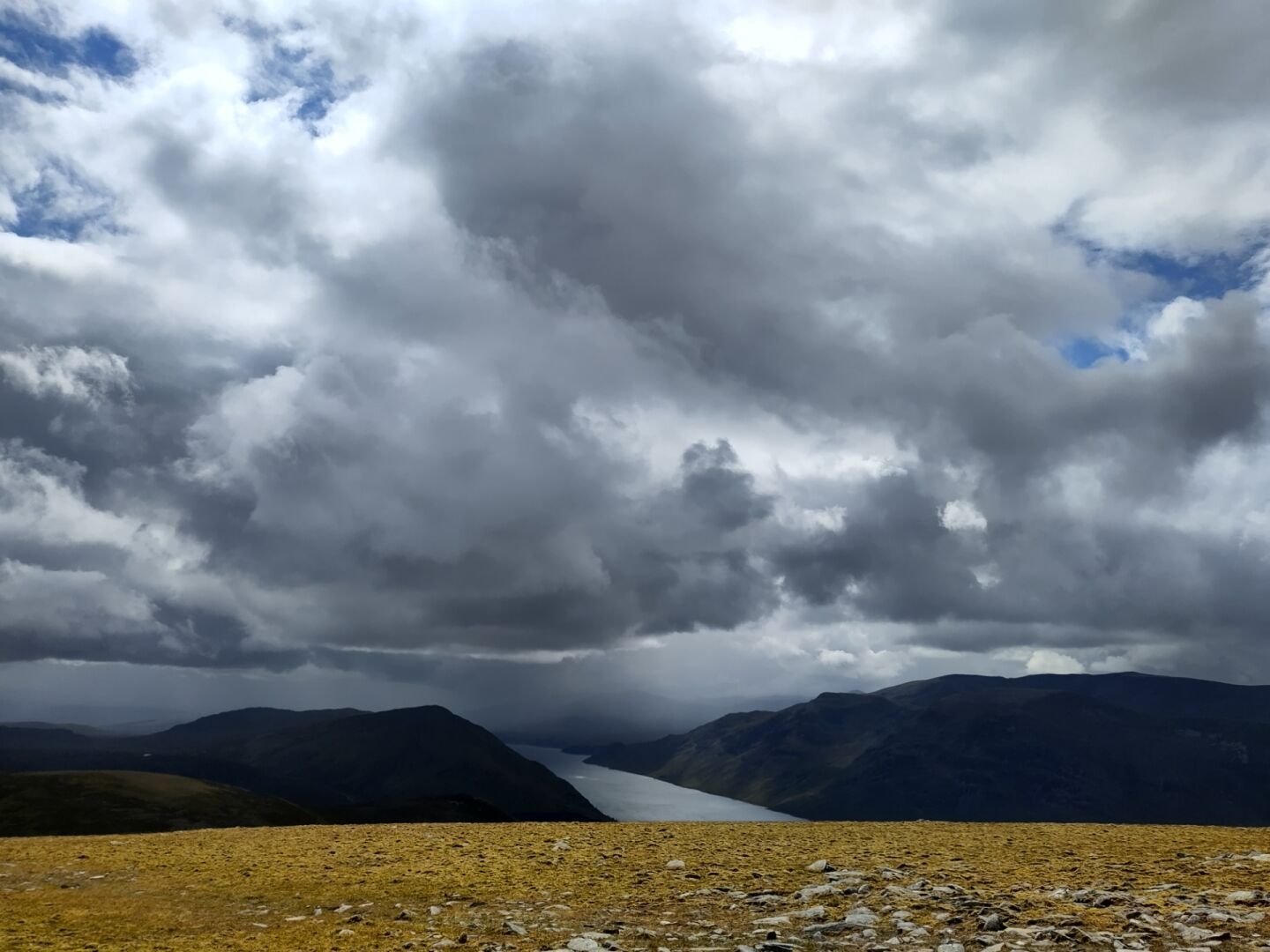 A threateningly dark sky full of big dark grey clouds with here and there bright blue. In the back grou d a long narrow lake stretches between two dark mountains.