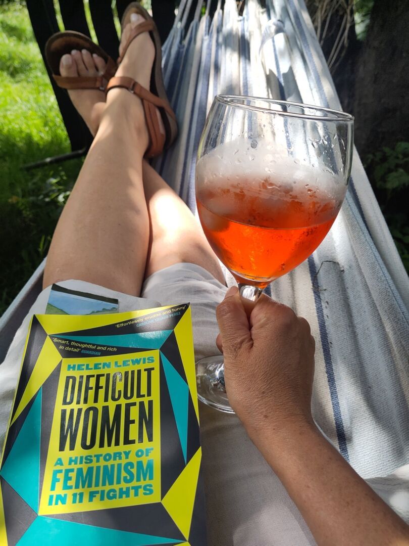 View from a hammock of legs, a hand holding a large glass with ice and an orange drink and a green and turquoise coloured book called Difficult women by Helen Lewis