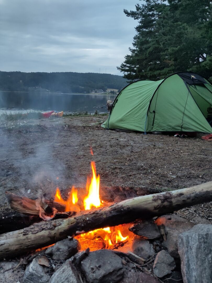 A campfire on a beach with a green tent in the background and a still fjord with forested hills in the background