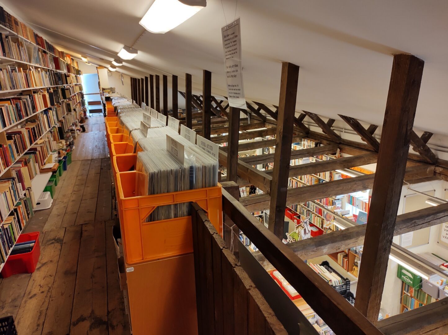 Inside of a used books store, looking down from the first floor to the ground floor. A row of orange plastic beer boxes filled with LPs in the foreground, heavy old beans are visible, it's an old building. The ground floor is filled with book shelves.