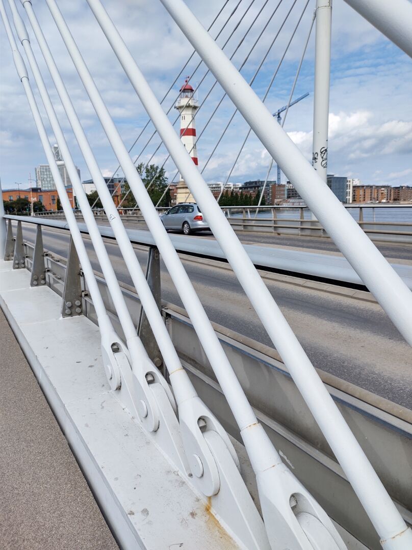 A white and red lighthouse seen through the crossed cables on either side of a bridge in the harbour.