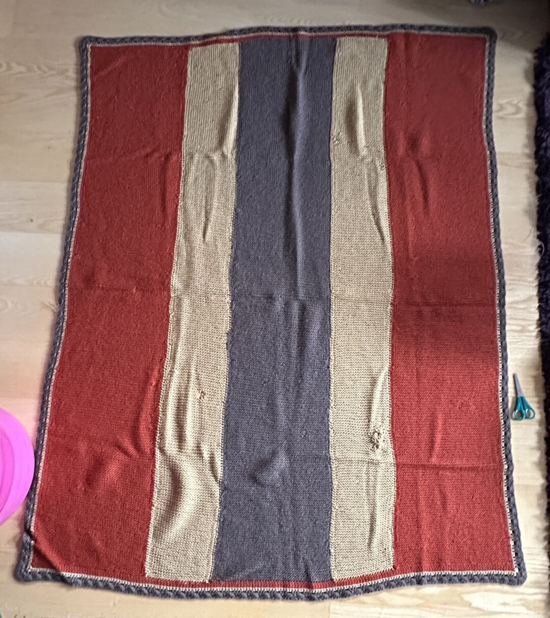 A garter stitch knitted blanket laid out on a wooden floor. The blanket has five wide vertical panels in colors rust, mustard, brown, mustard and rust, and a brown crochet border. In the middle of the left rust panel is a hole, a little further down in the adjoining mustard panel, too. The brown panel looks okay, just some thin spots. The right mustard panel has a big hole which was mended very inexpertly, but at least they tried.