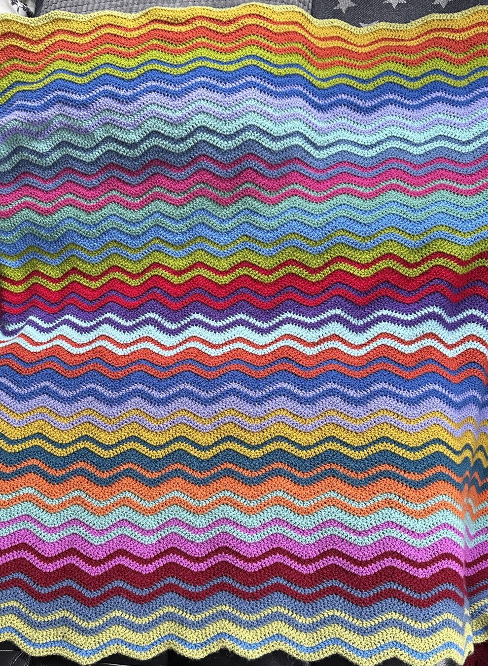 Laid out Ccrochet blanket in progress. The pattern are interlocking ripples in colours all over the rainbow. It looks bright and happy.