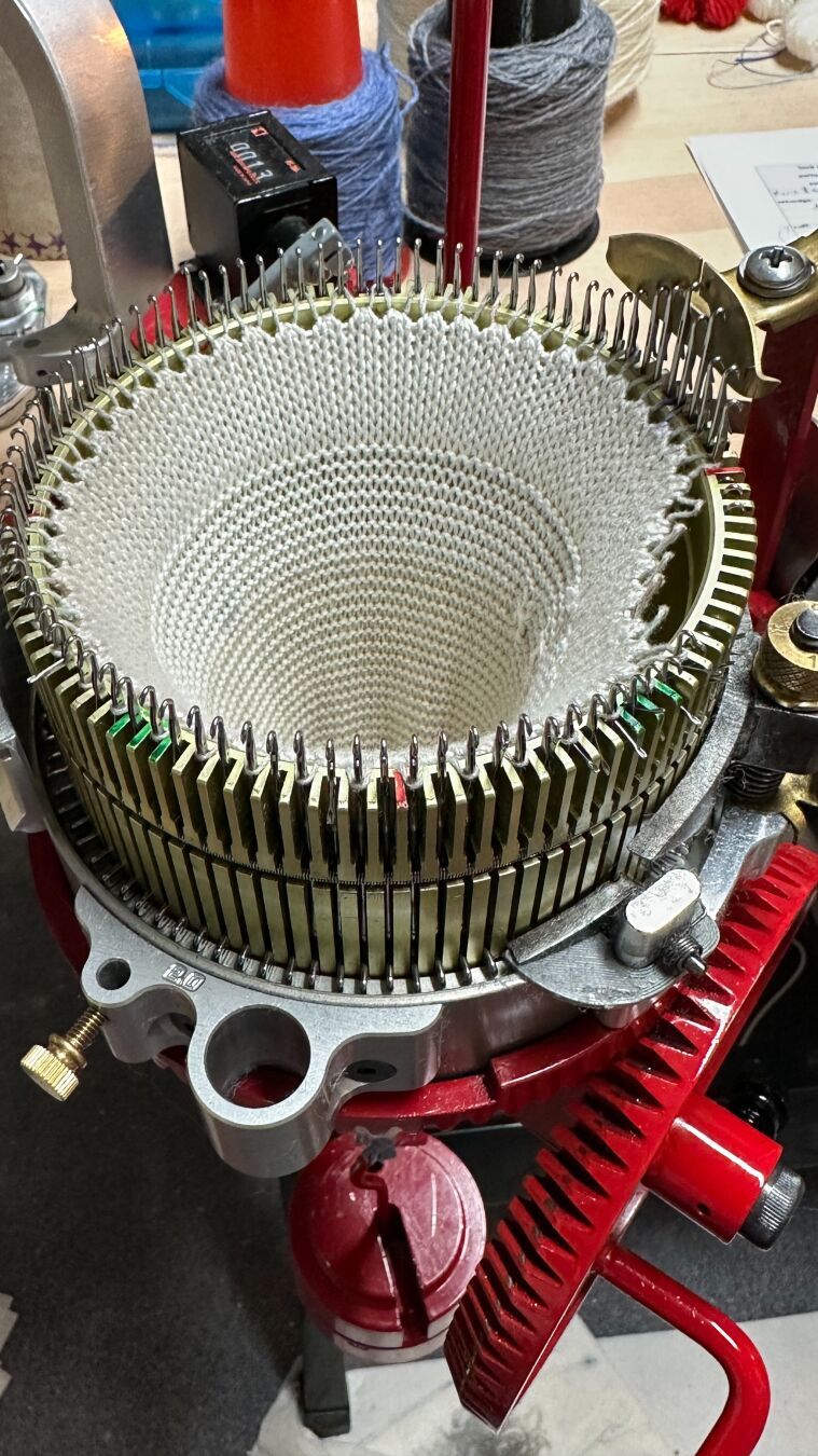 View from slightly above of a CSM. It‘s a red Erlbach Gearhart with a 96 needle cylinder. It is set up to start a project, with the cast-on bonnet already hung on every other needle up to the last few needles. Start of rounds is at 3 o‘clock.