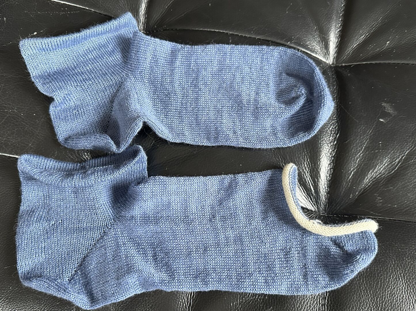 Two blue sneaker socks laid out on a black leather background. The sock on the top is done, the sock on the bottom still needs to be sewn closed across the toes, there‘s still white divider yarn attached to the live stitches.
