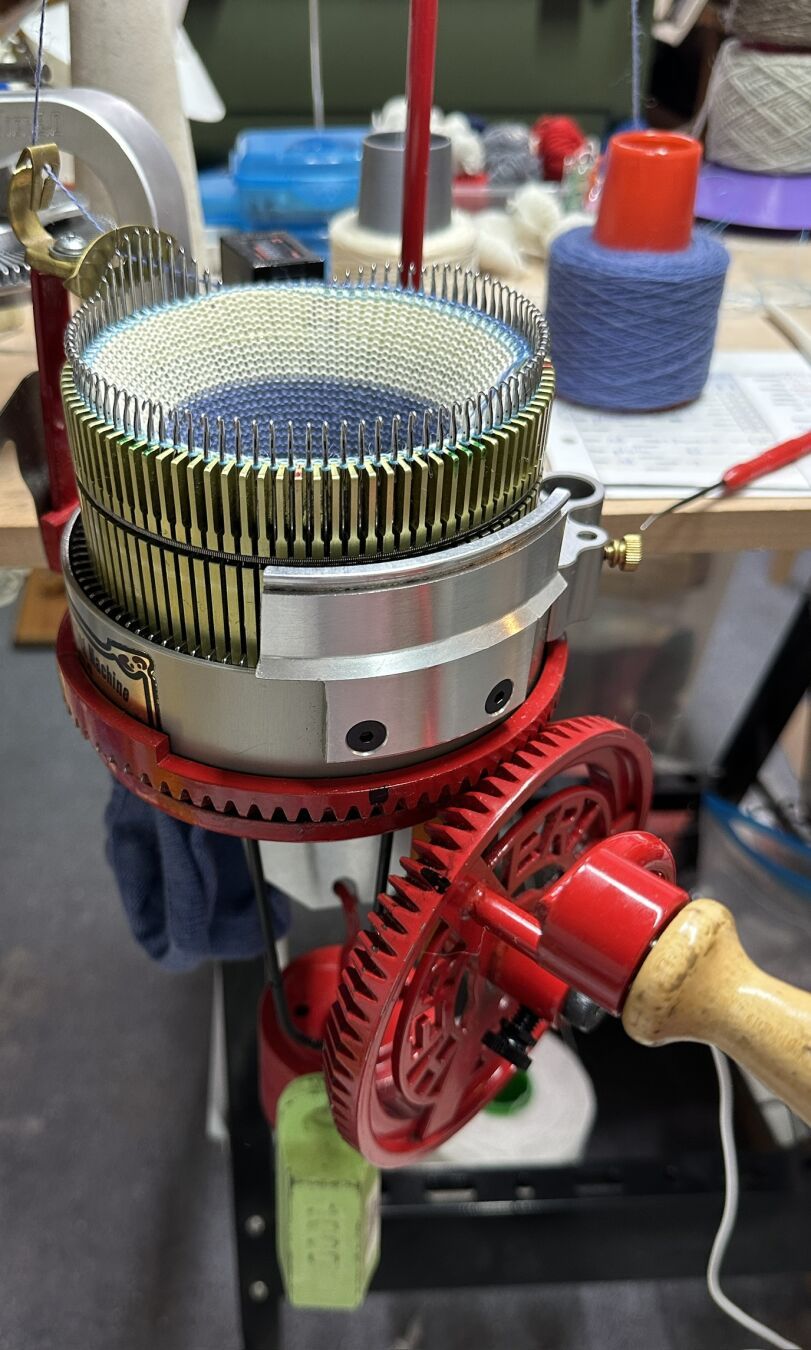 A red hand-crank circular sock machine with a 96-needle cylinder. On the machine is a work in progress, a pair of blue sneaker socks. The first sock is done already and can be seen under the machine with weights hanging from it. The knitted fabric close to the needles is white divider yarn, and the last knitted row is light blue fishing line as ravel cord.
The machine is attached to a small work bench with cones of yarn, boxes with needles and some tools on it.