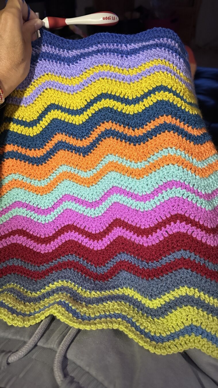 An in-progress crochet blanket with interlocking ripple stripes photographed laid out on a person‘s lap. Stripe colors from bottom to top are so far: Pistachio, Denim, Claret, Magenta, Spearmint, Petrol, Mustard, Lavender, and Lapis.