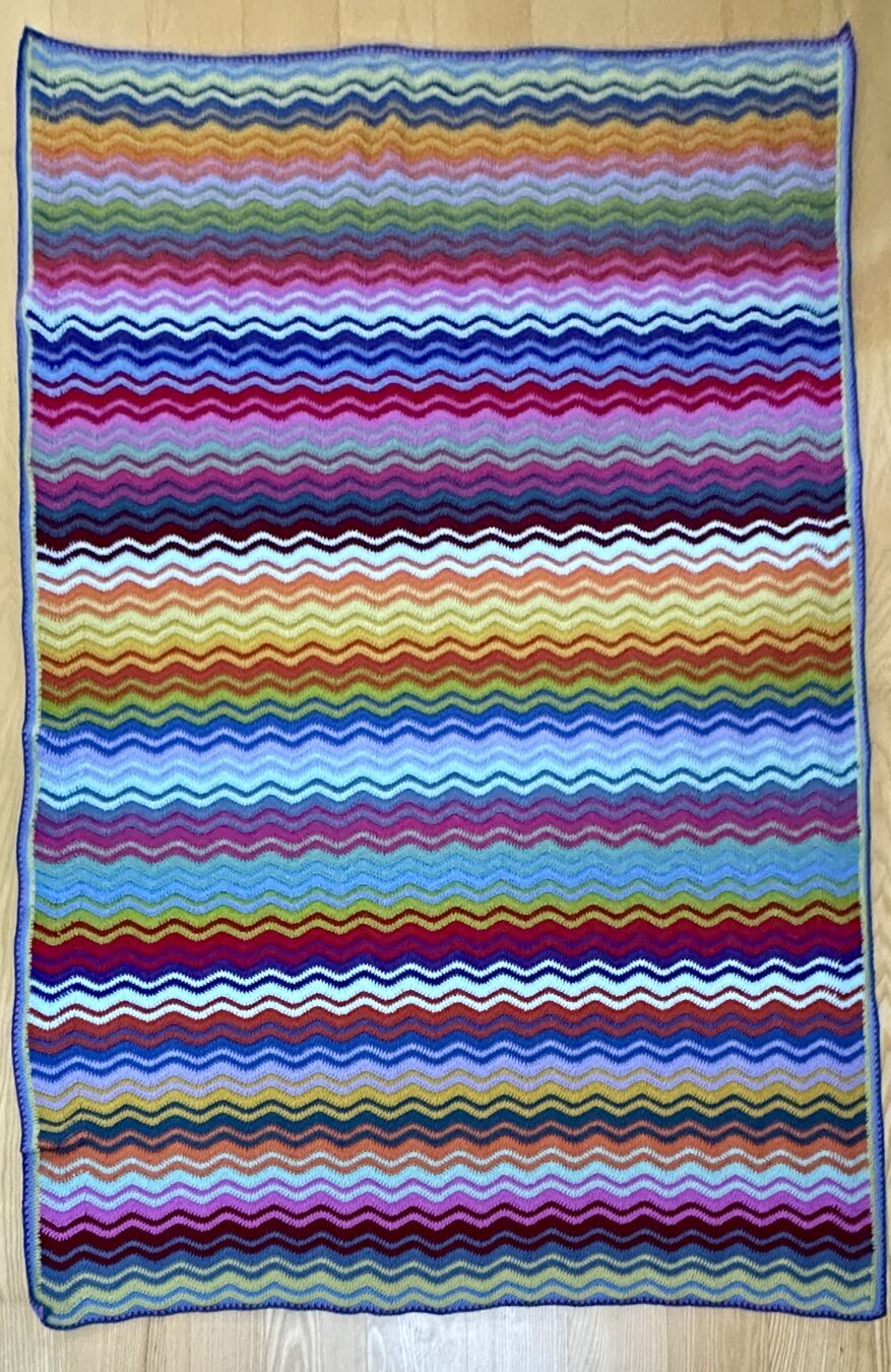 The full blanket, laid out flat on the floor. All 18 colors in random interlocking ripple stripes are visible.